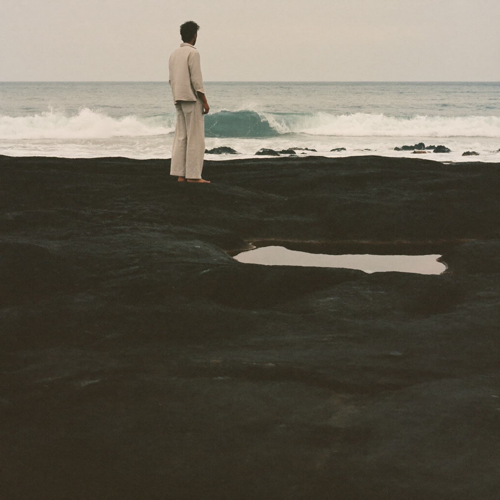 A man alone, seen from behind, standing on the black basalt of the Atlantic shoreline in Casablanca facing the swell, in the light of a hazy morning